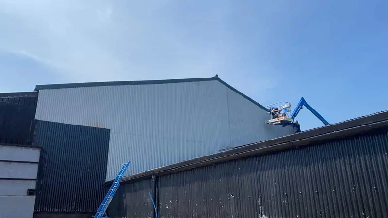 Operatives in a boom lift at the eaves of a large industrial building clad in light grey profiled metal sheeting