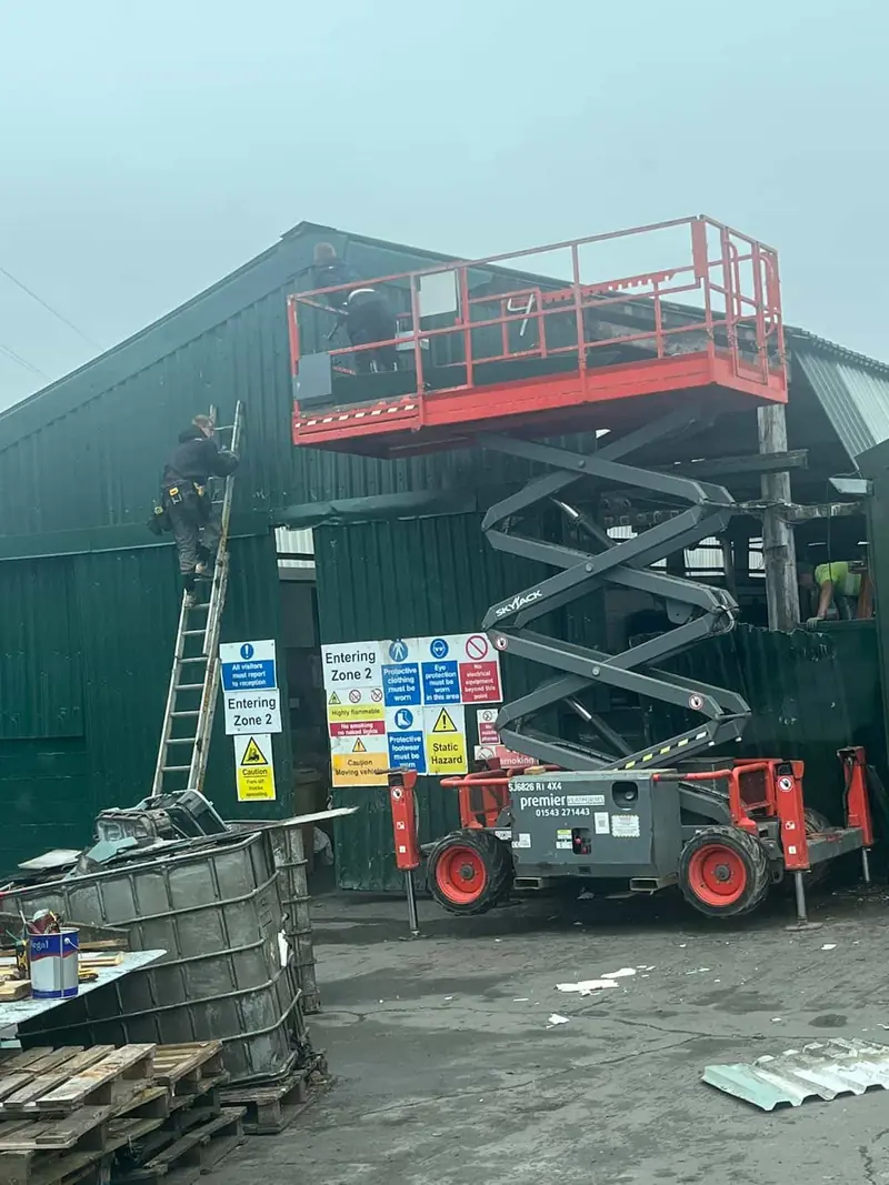 Operatives on a scissor lift and ladder re-cladding a green steel-clad industrial building on a working site