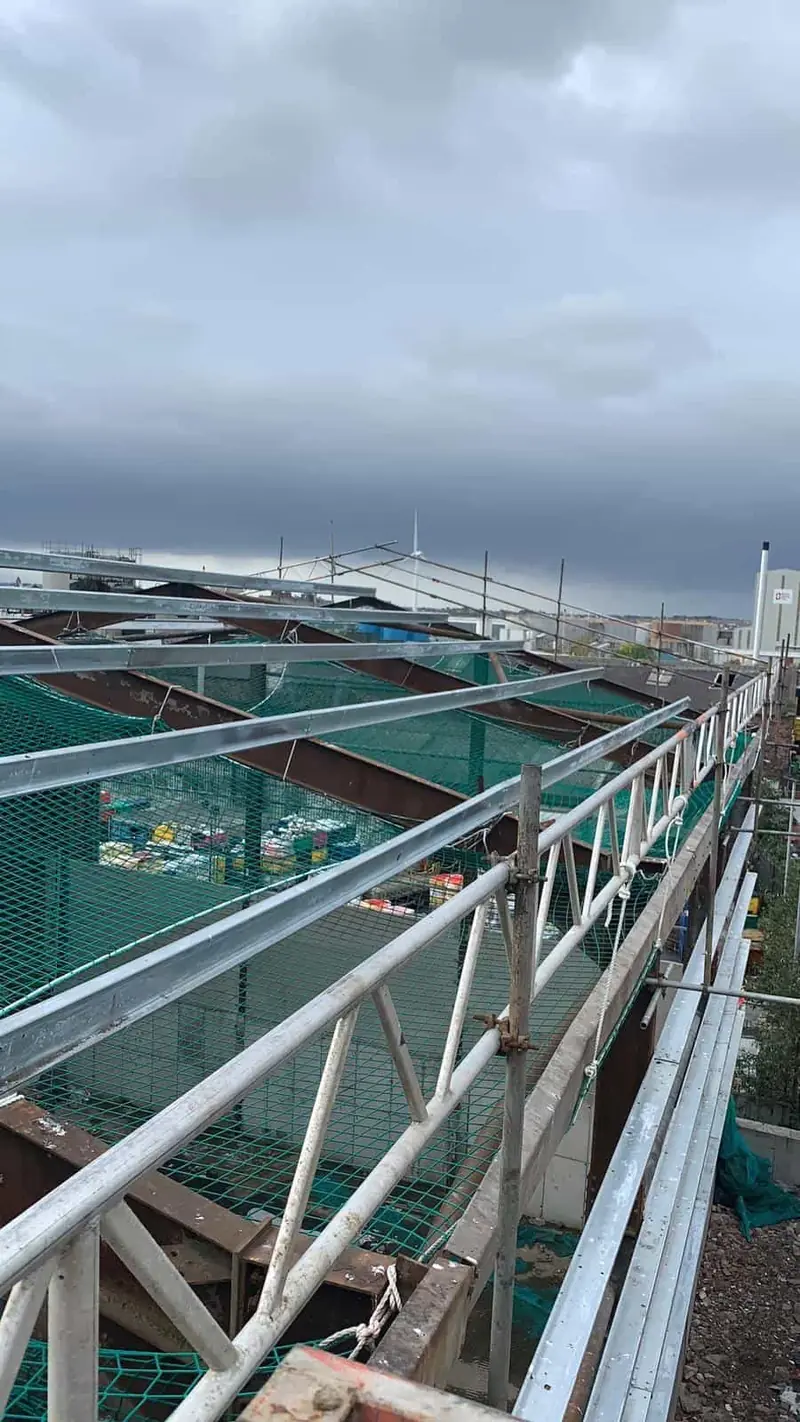 Stripped industrial roof with safety netting across the bays and edge protection along the gutter line