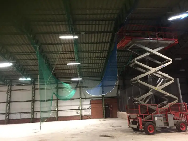 Inside a warehouse, with safety netting rigged beneath the roof and a scissor lift in position for roof works